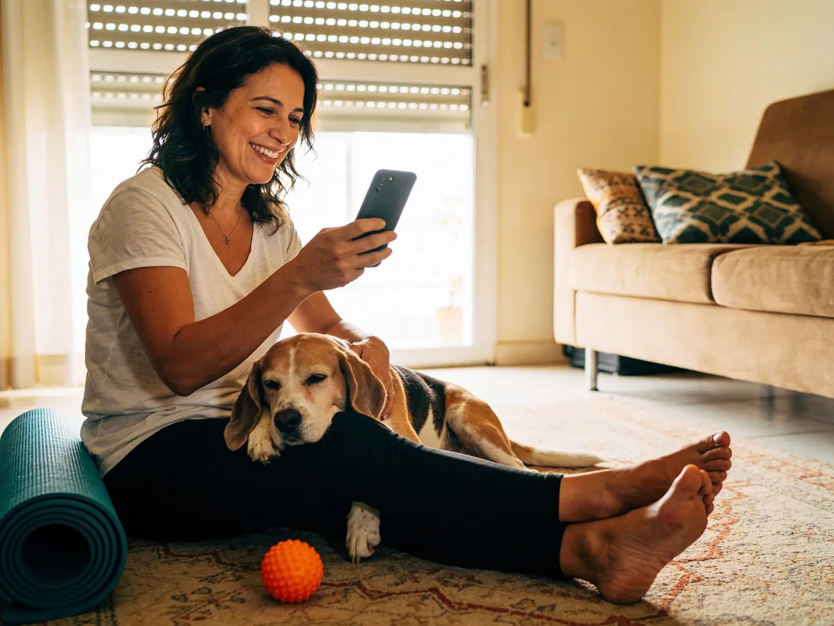 Responsável sorrindo em casa, sentada no tapete com o beagle no colo, olhando o app no celular.